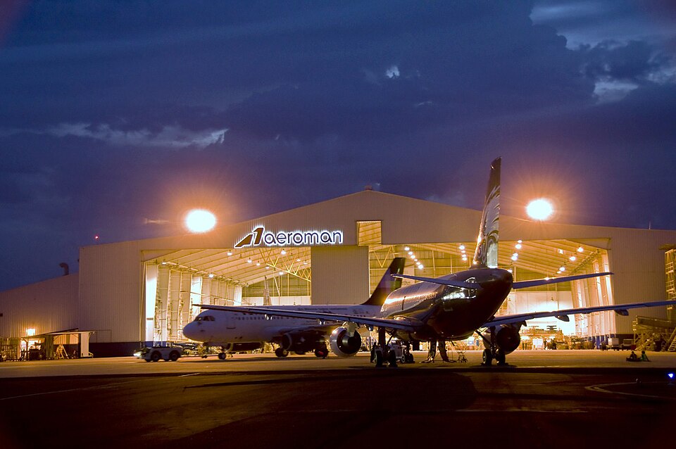 An airliner on the apron in front of the lit Aeroman maintenance hangar in El Salvador at dusk, photographed in June 2010
