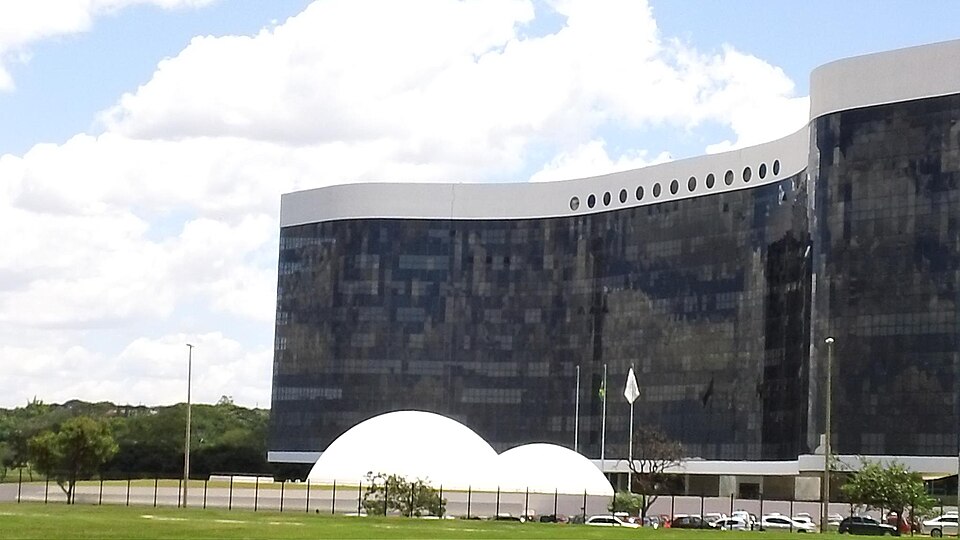 The Superior Electoral Court's headquarters in Brasília: a long, curved building of dark reflective glass with a white rim and a row of round windows along its top, two white domes in front, flagpoles