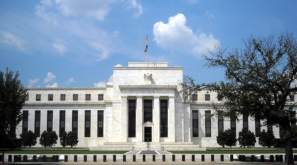 The white marble front of the Marriner S. Eccles Federal Reserve Board Building in Washington, with an eagle above the entrance and a US flag on the roof under a blue sky
