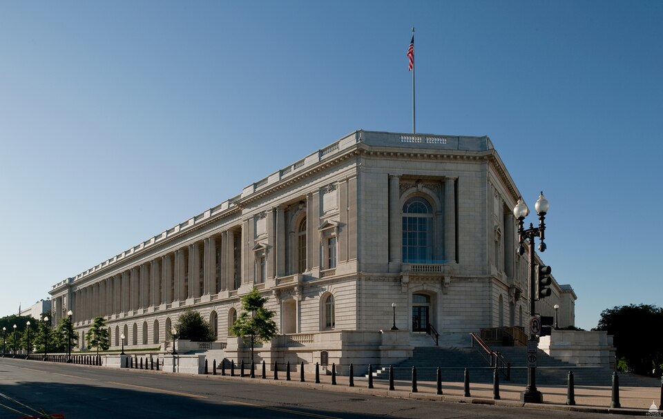 The Cannon House Office Building of the House of Representatives in Washington, a long white neoclassical building with an American flag on the roof, under a clear blue sky