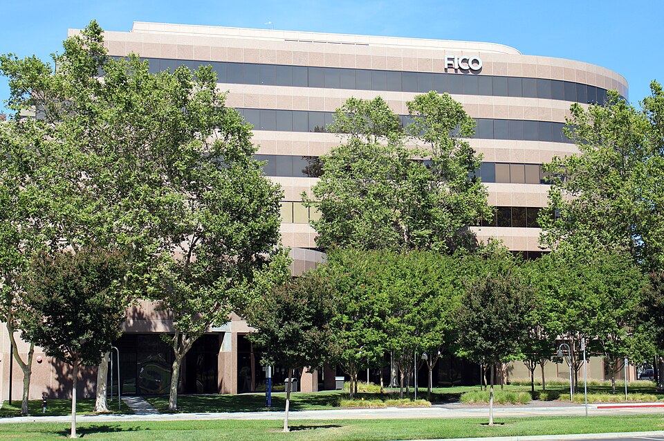 A curved beige office building with FICO lettering near the roof, behind a row of green trees under a blue sky, photographed in San Jose, California in June 2020