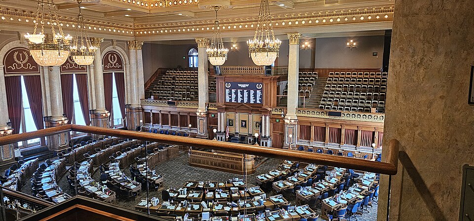 The chamber of the Iowa House of Representatives in the State Capitol in Des Moines, seen from behind the gallery rail in March 2025: curved rows of members' desks face a raised dais at the front, ben