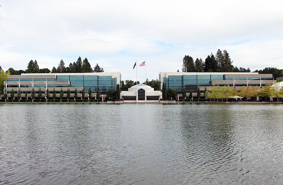 Two glass-fronted office buildings at Nike's world headquarters in Beaverton, Oregon, seen across a lake, with a small white entrance building and two flags between them, in September 2017