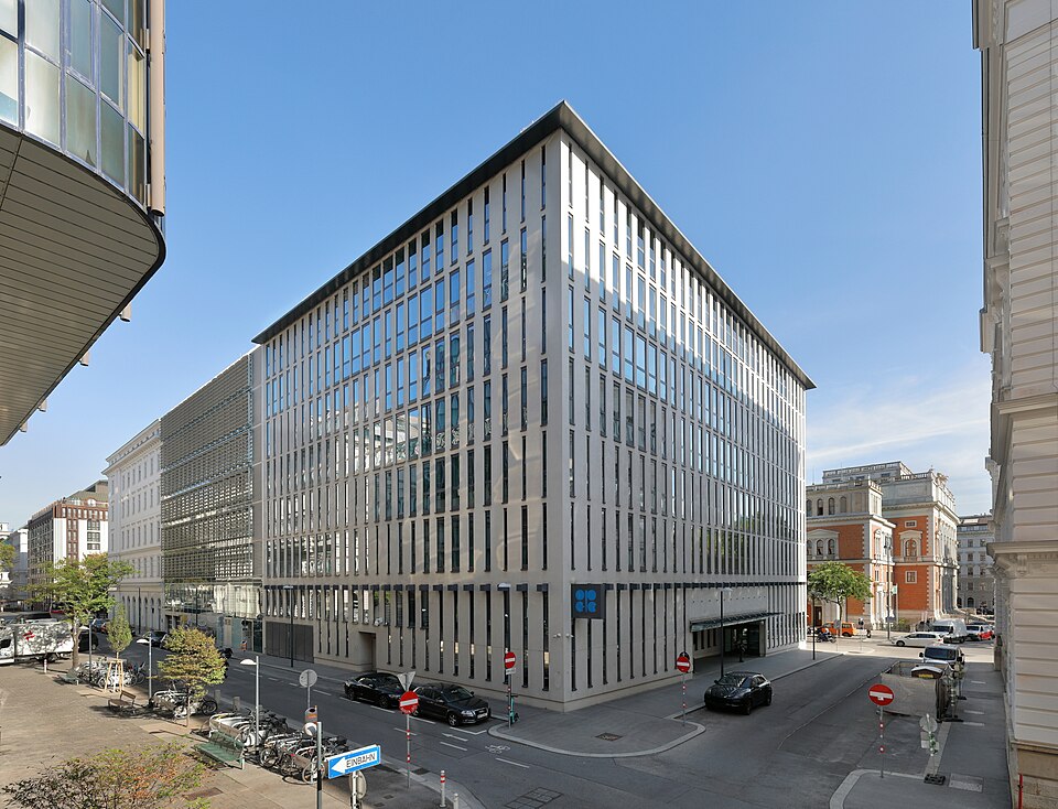 The OPEC headquarters in Vienna, a pale multi-storey office block with narrow vertical windows and the blue OPEC emblem at its corner, seen from a sunny street with parked cars and bicycles