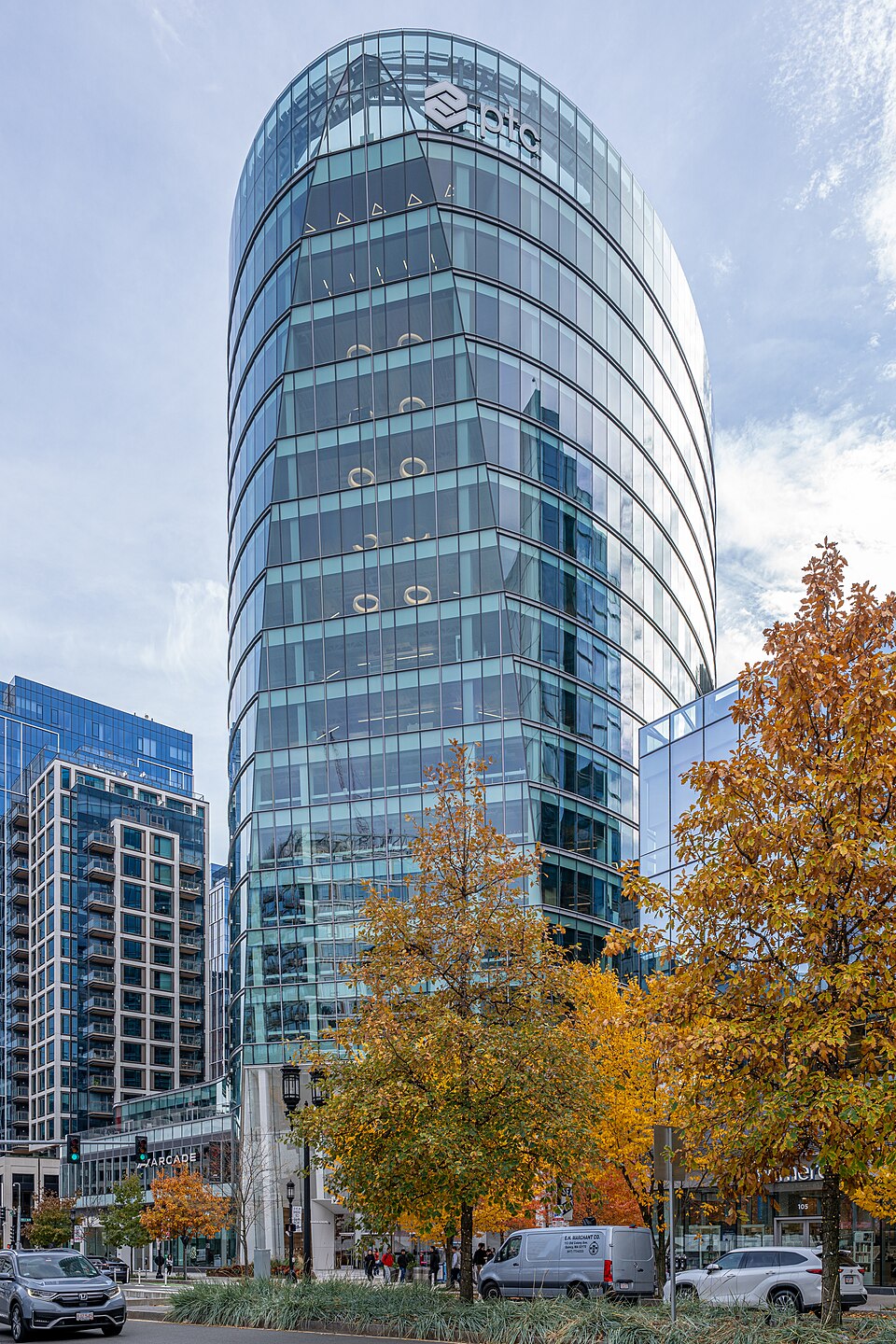 A curved glass office tower with the PTC name and logo near its top, seen from a Boston street lined with autumn trees, with a van and cars passing in front