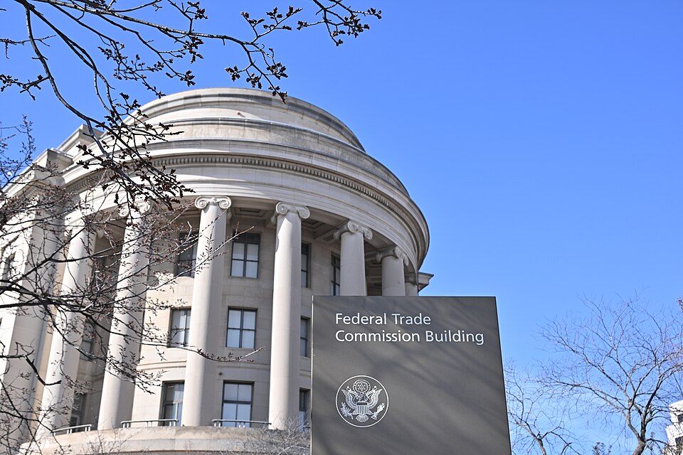 The curved, columned end of the Federal Trade Commission's headquarters in Washington behind bare winter branches, with a sign reading Federal Trade Commission Building in front, under a clear blue sk