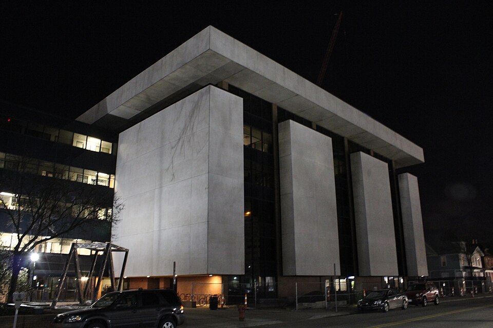 The Institute for Social Research building of the University of Michigan in Ann Arbor at night, tall pale stone panels under a wide overhanging roof, with cars parked along the street in front