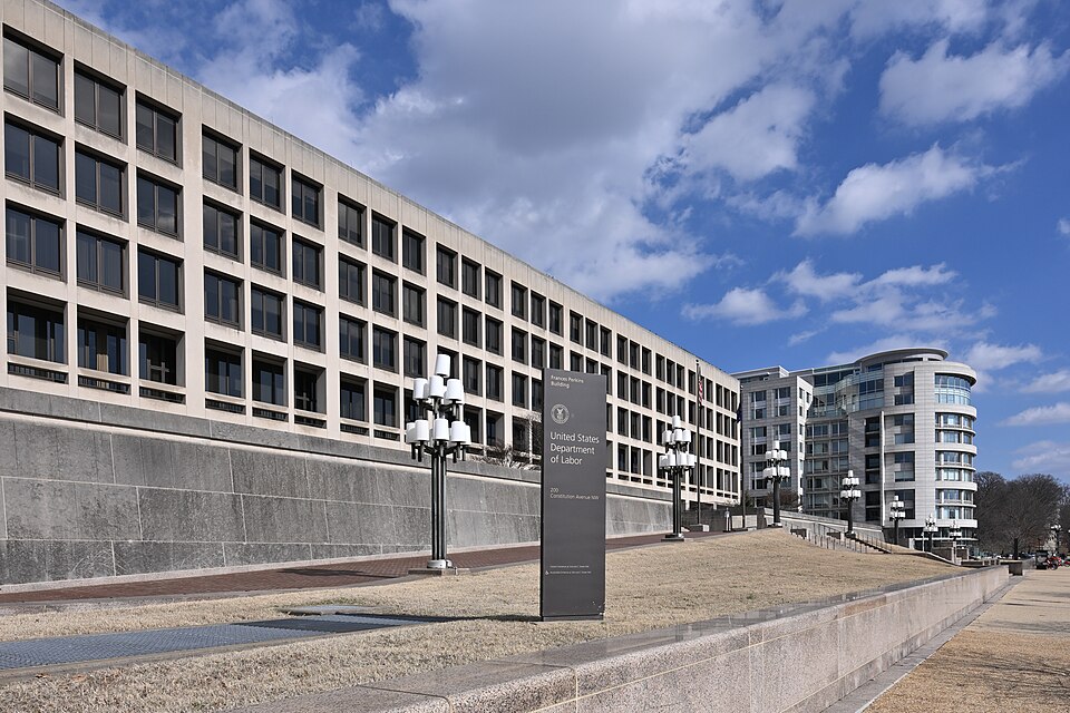 The sign of the United States Department of Labor on Constitution Avenue in Washington, with the Frances Perkins Building behind it under a blue sky, in February 2025