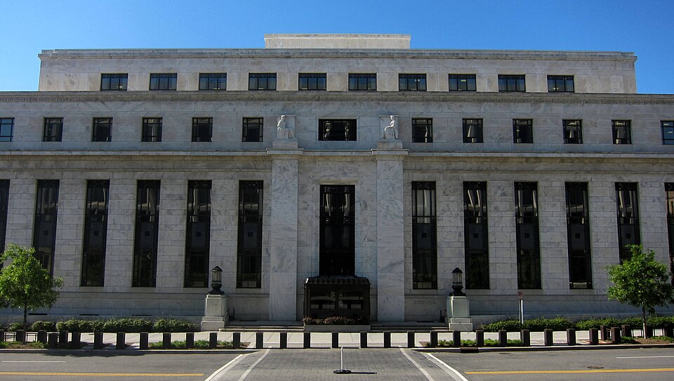 The north side of the Marriner S. Eccles Federal Reserve Board Building in Washington, D.C., a pale marble facade with tall dark windows and two carved figures over the entrance, under a clear blue sk