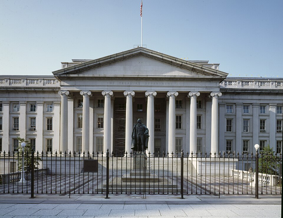 The columned front of the U.S. Treasury Department building in Washington, D.C., with 'The Treasury Department' carved above the columns, a flag on the roof, a bronze statue of a standing figure befor