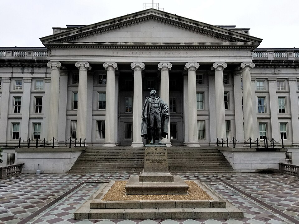 The north entrance of the U.S. Treasury Building in Washington, D.C., a columned portico, with the bronze statue of Albert Gallatin in the plaza in front, under a grey sky