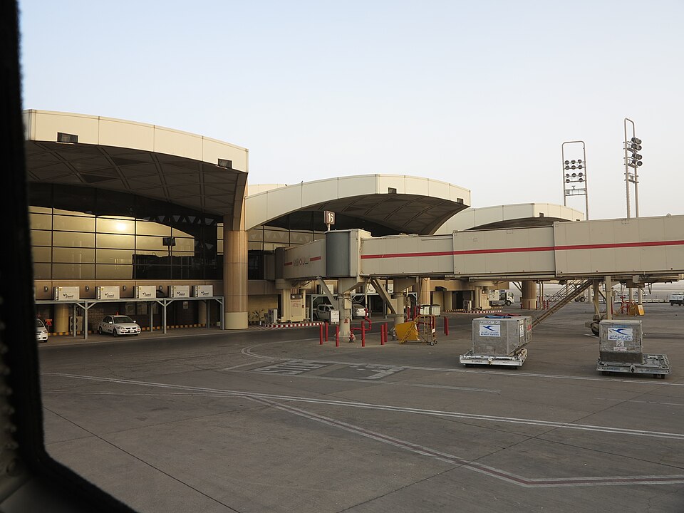 Terminal 1 at King Khalid International Airport in Riyadh, seen from an aircraft window: a curved concourse with a tinted glass front, a jet bridge at gate 16 and cargo containers waiting on the apron