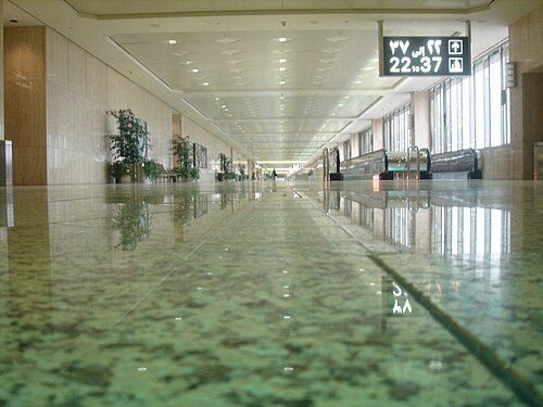A long, empty concourse at King Fahd International Airport in Dammam, photographed from floor level: a polished green and white granite floor reflects ceiling lights, potted plants, rows of seats alon