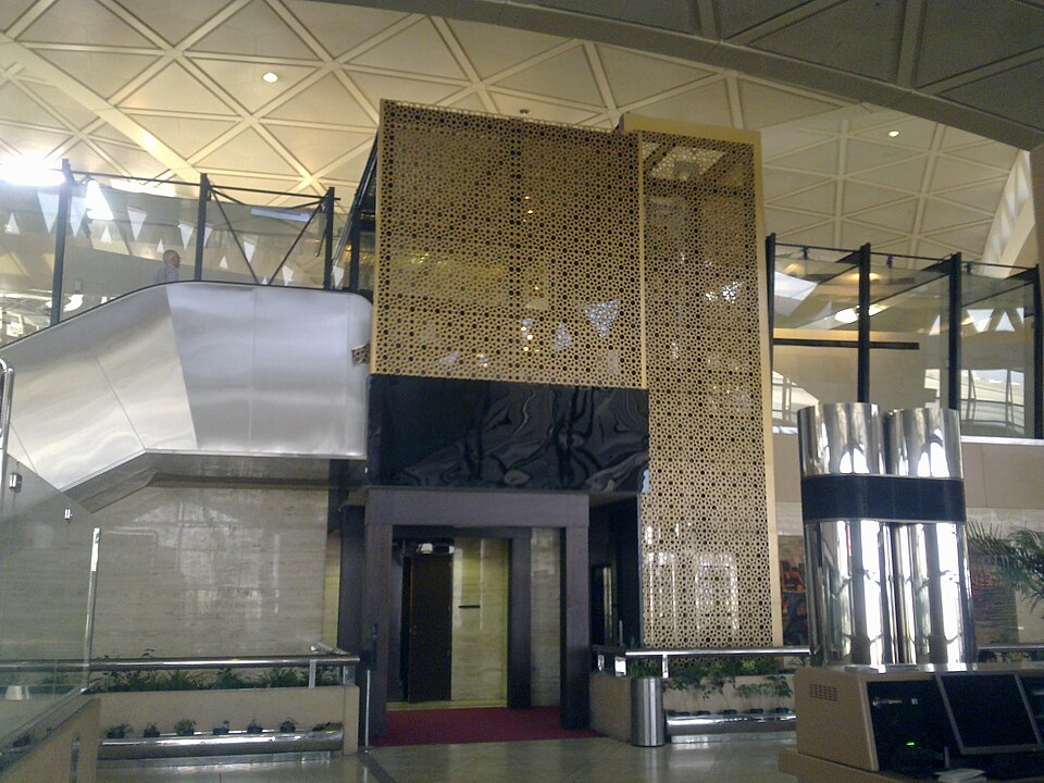 Inside a terminal at King Khalid International Airport in Riyadh: a lift shaft clad in gold lattice panels rises under a white ceiling of triangular panels, beside a steel staircase and glass balconie