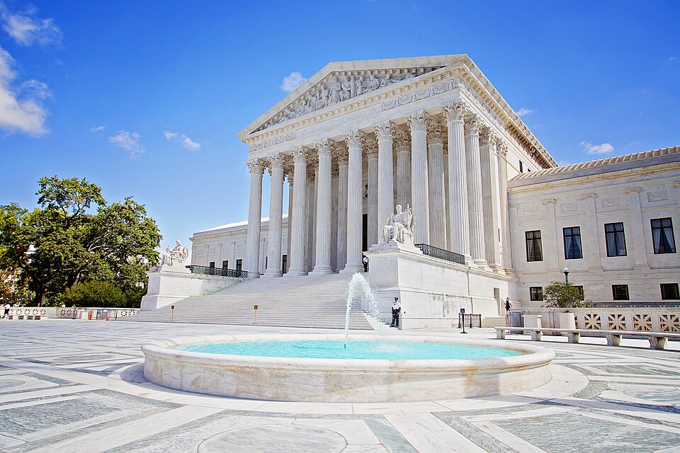 The Supreme Court Building in Washington under a blue sky: the white marble front with its columns and sculpted pediment above wide entrance steps flanked by two seated statues, and a fountain in the 