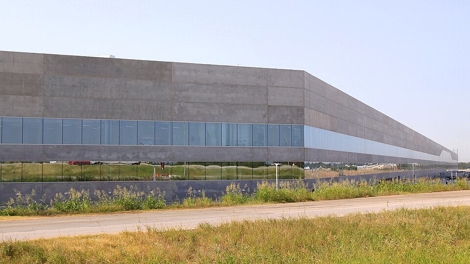 Building 1 of Tesla's Gigafactory Texas near Austin, a long grey concrete building with a band of windows, behind a field of grass in June 2022