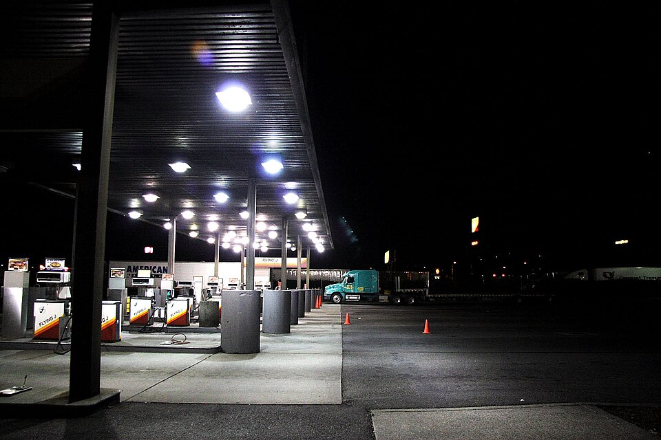 Diesel pumps under a lit canopy at a truck stop at night, with a semi truck parked beyond them