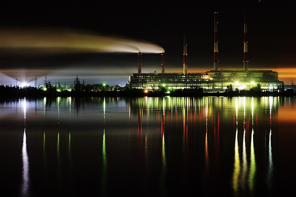 A thermal power plant with tall striped chimneys lit up at night, its lights reflected in a reservoir in the foreground; the Commons file names it the Zmiiv thermal power plant in Ukraine, photographe