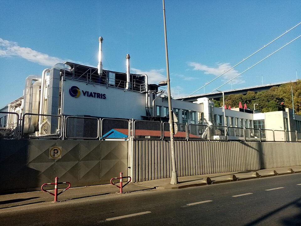 A white Viatris pharmaceutical plant with tall exhaust stacks and the company's name on its wall, behind a fence beside a road in Ortakoy, Istanbul, with a bridge's cables overhead
